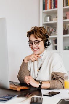 Smiling happy female in eyeglasses and wired headphones working on modern computer in contemporary light workspace in daytime