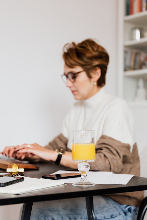 Side view pensive concentrated blurred female in trendy wear typing on computer keyboard while working at desk with papers and glass of fresh orange juice