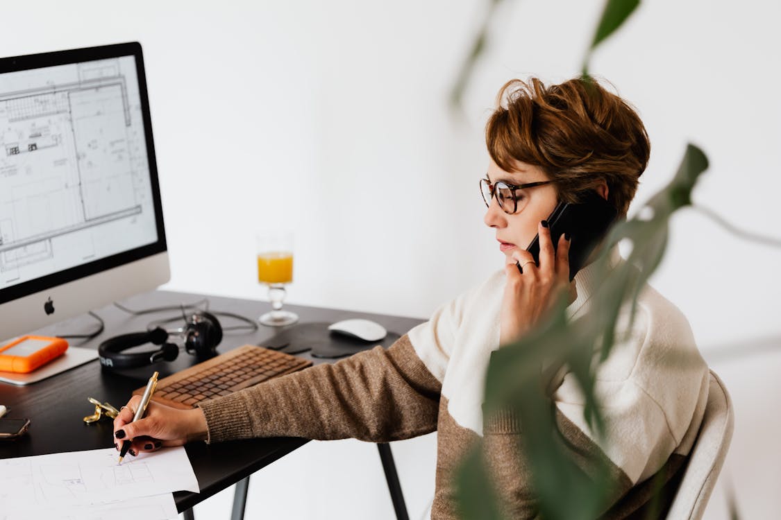 Side view of serious female specialist in casual clothes and eyeglasses checking information via smartphone and taking notes on paper