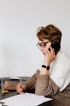 A focused businesswoman in an office environment, working and speaking on a smartphone.