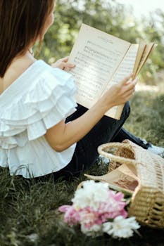 Woman sitting on grass reading music sheets outdoors, embodying a serene and artistic vibe.