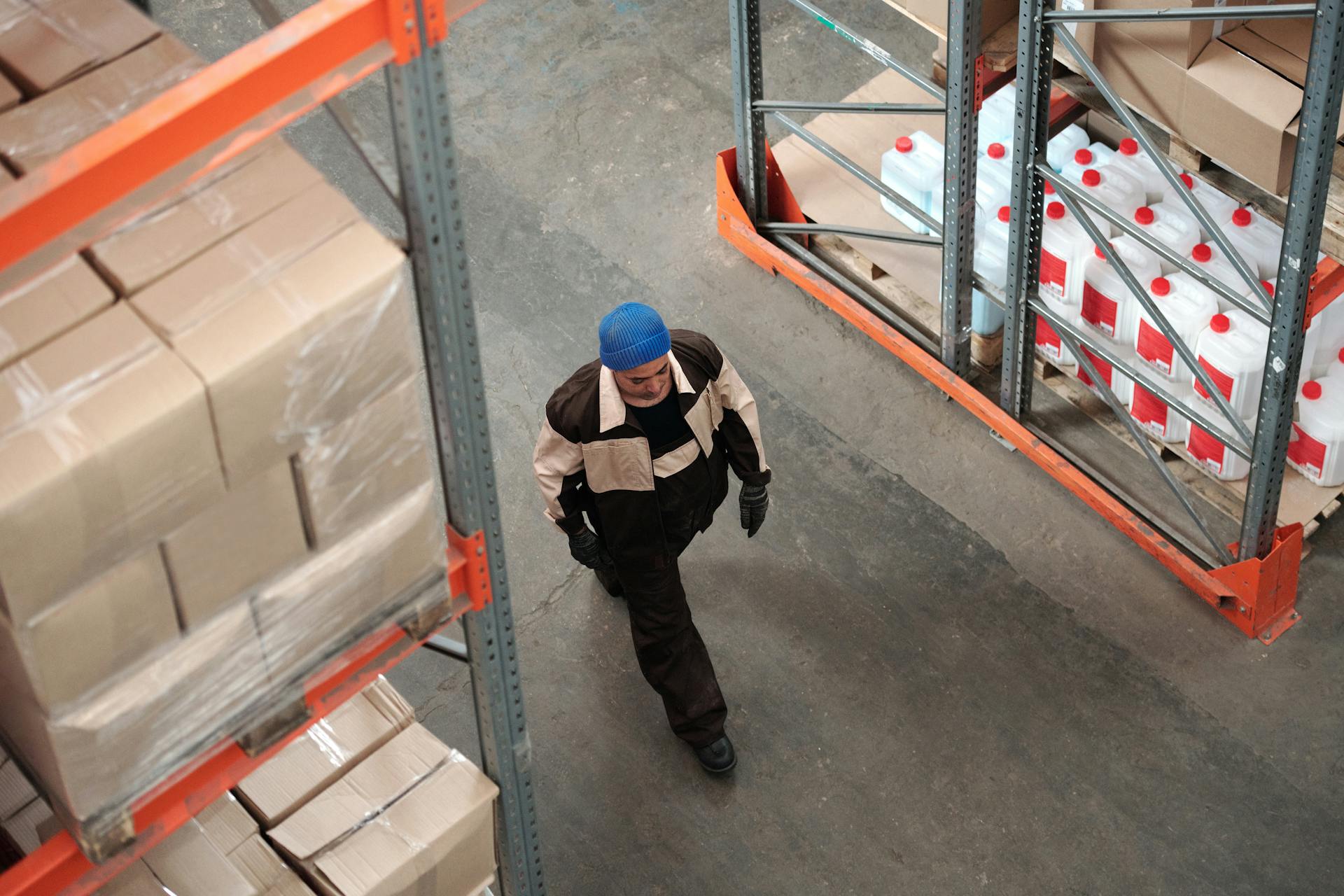 A warehouse worker in a blue hat walking past stacked packages and shelves in an industrial setting.