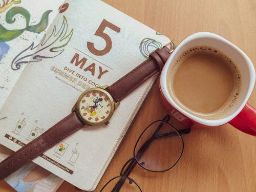 Top view of cup of fresh coffee and notepad arranged on table with wristwatch and eyeglasses in creative workspace of freelancer