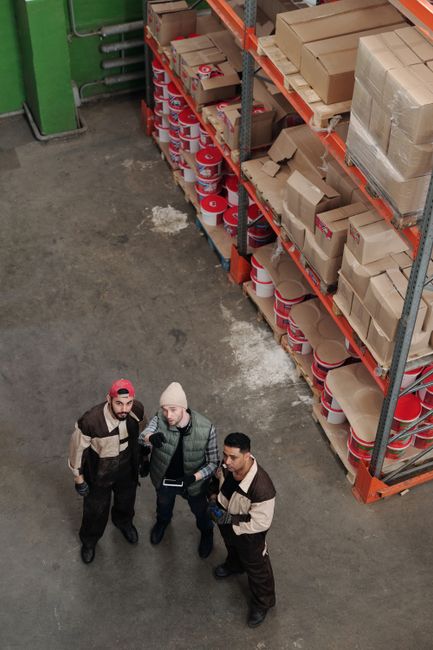 Workers in a warehouse discussing logistics near shelves filled with packages and products. Workers in a warehouse discussing logistics near shelves filled with packages and products.