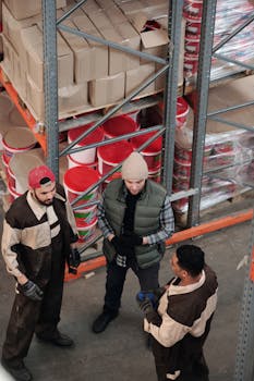 Group of warehouse workers discussing logistics in an industrial storage area with shelves and packaging.
