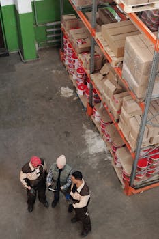 Group of warehouse workers discussing logistics and inventory in a large storage area with shelves and packages.