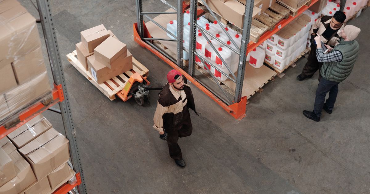 Mitarbeitende in einem Logistiklager bewegen Paletten zwischen Regalen, laufender Betrieb während der Reinigungsplanung