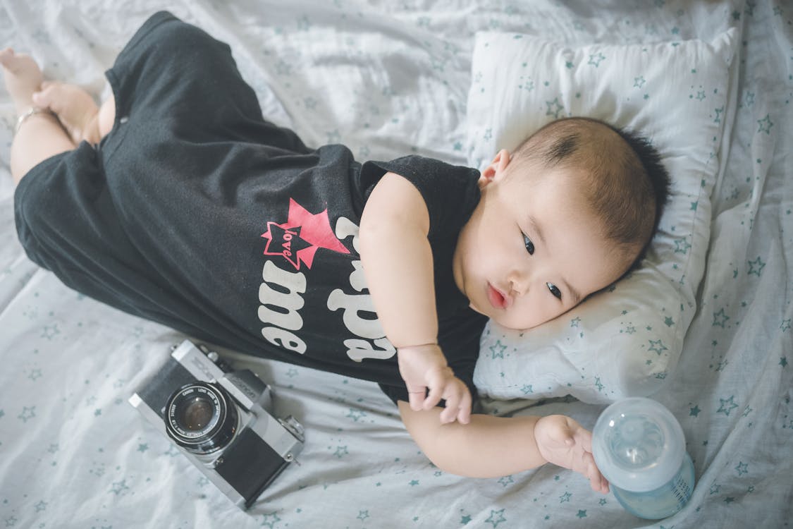 From above of cute Asian baby boy in bodysuit reaching out for bottle with milk while lying on soft bed with photo camera placed nearby