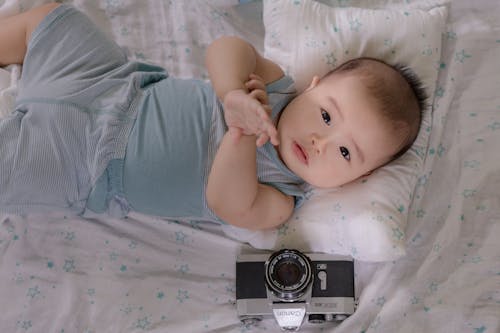 Free Cute ethnic infant lying on bed with photo camera Stock Photo