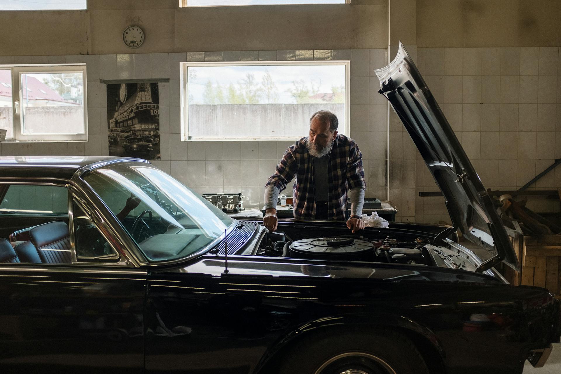 A bearded mechanic examines a classic car engine in a well-lit auto repair shop.