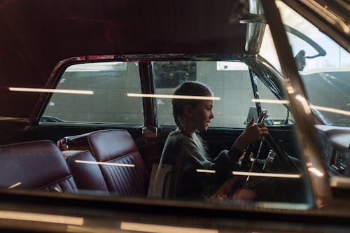Free A young girl with pigtails sits in a vintage car, holding the steering wheel with focus. Stock Photo