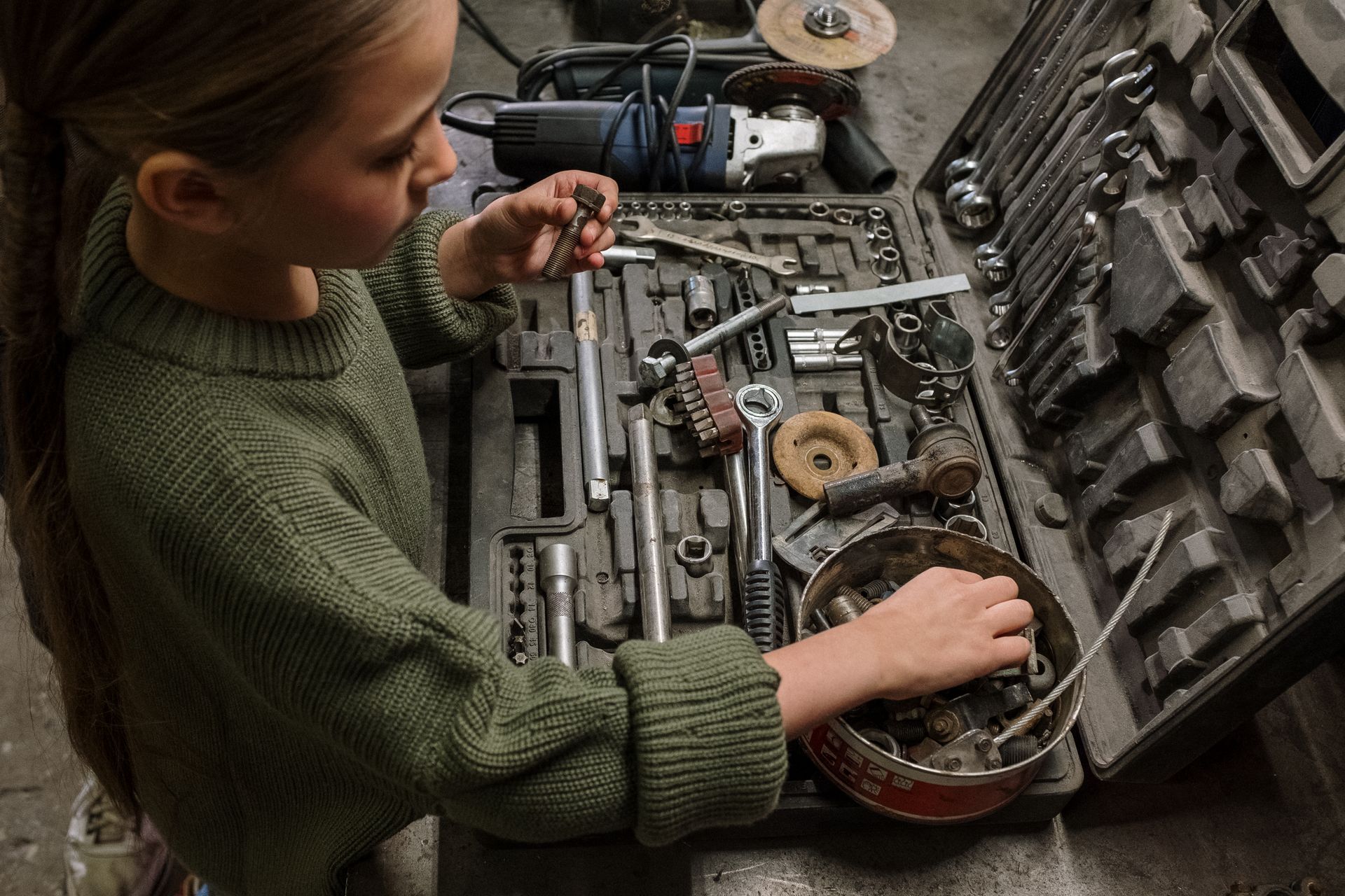 A young girl examines assorted tools and equipment in a garage workshop, depicting curiosity and hands-on learning.