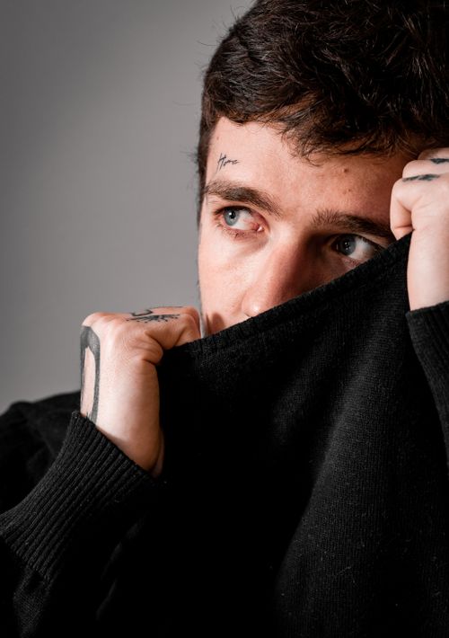 Free A thoughtful man with tattoos looks away, partially covering his face with a black sweater. Stock Photo