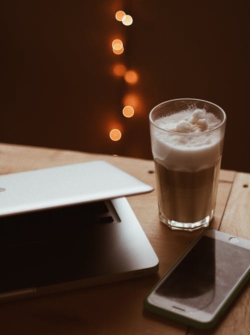 Close-Up Photo of a Foamy Drink in a Glass beside a Smartphone and Laptop
