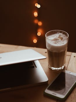 A warm-toned image showing a glass of frothy coffee beside a laptop and smartphone with bokeh lights.