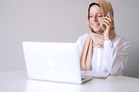 一位戴着头巾的妇女微笑着在家庭办公室里使用笔记本电脑和电话