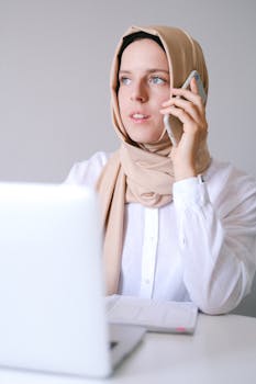 Muslim woman wearing hijab working remotely while talking on smartphone in home office.