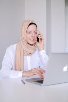 A woman wearing a hijab uses a laptop and phone for remote work, smiling in a home office.