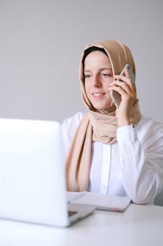A modern Muslim woman in a hijab using a smartphone and laptop for remote work.
