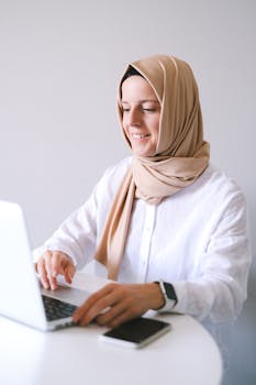 A smiling Muslim woman in a hijab working on a laptop from a home office.