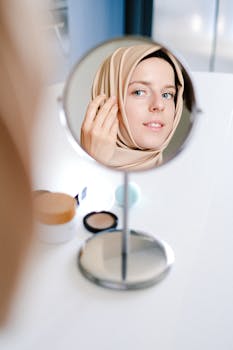 A woman in hijab applying makeup at a mirror, smiling and relaxed.