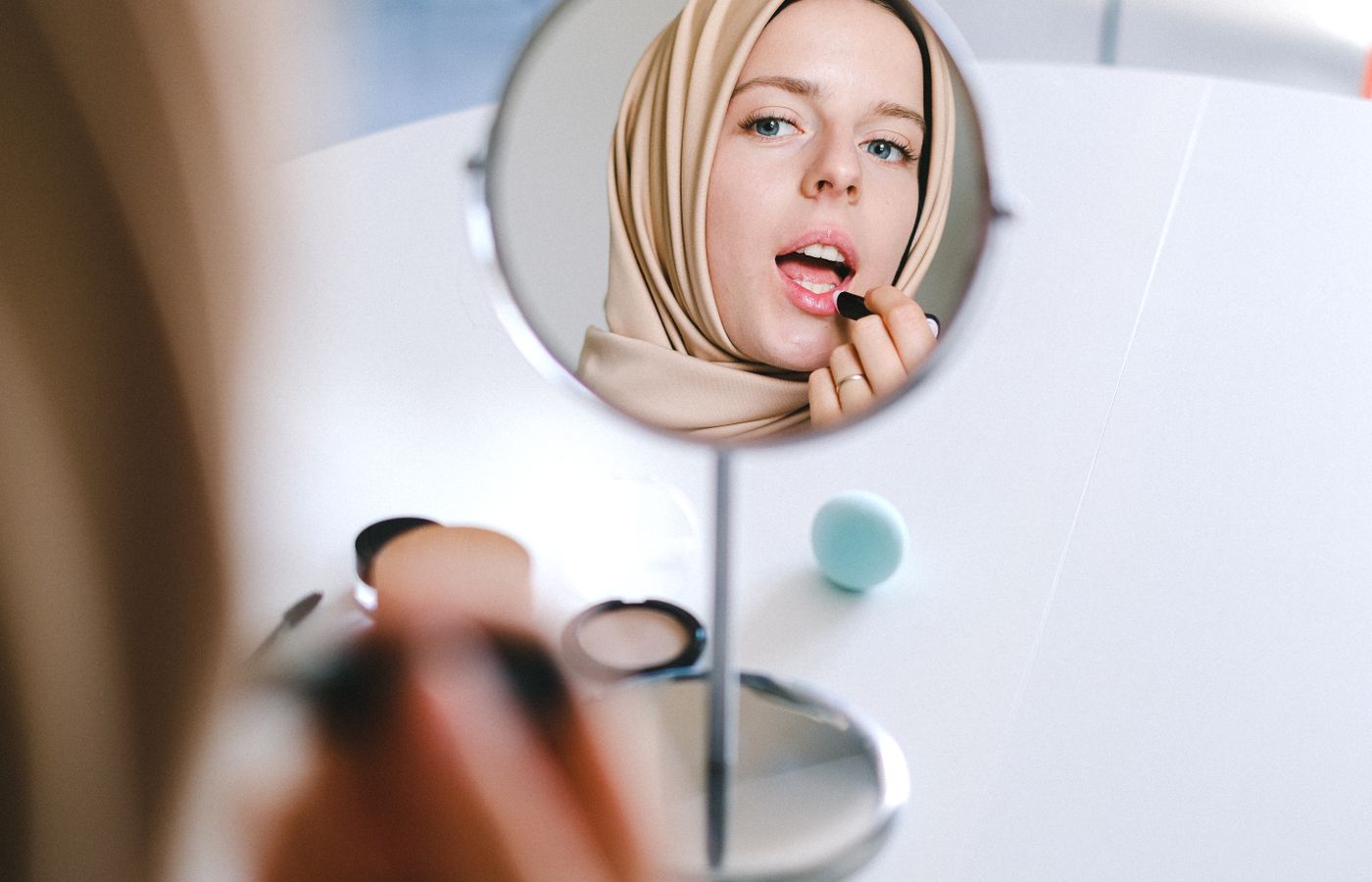 Muslim woman in hijab applies lipstick while looking at her reflection in a mirror.