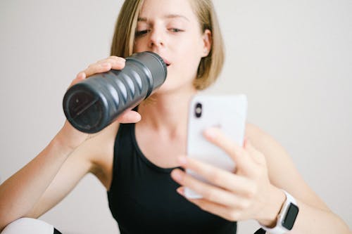 Free Woman in Black Tank Top Holding White Smartphone While Drinking Stock Photo