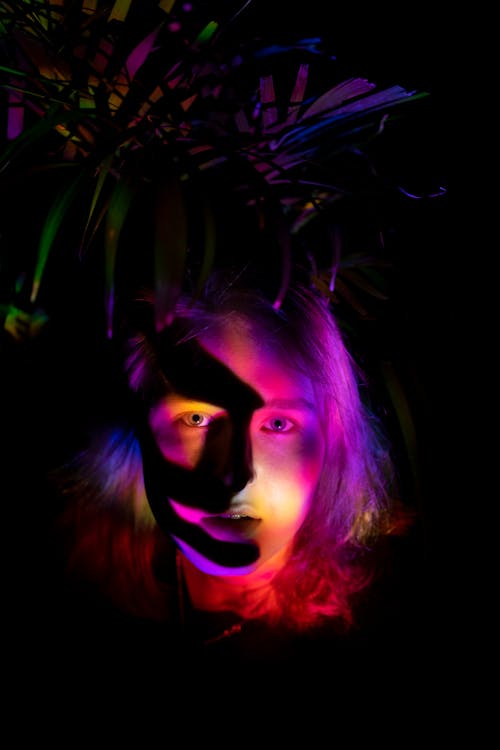 Young female illuminated by colorful neon lights with shadow on face standing in darkness against bush