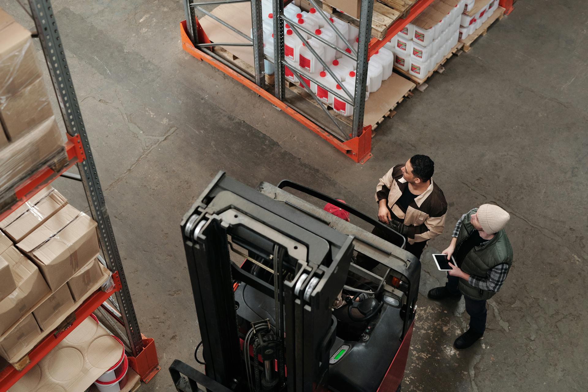 Two workers in a warehouse discussing logistics near a forklift captured from above.