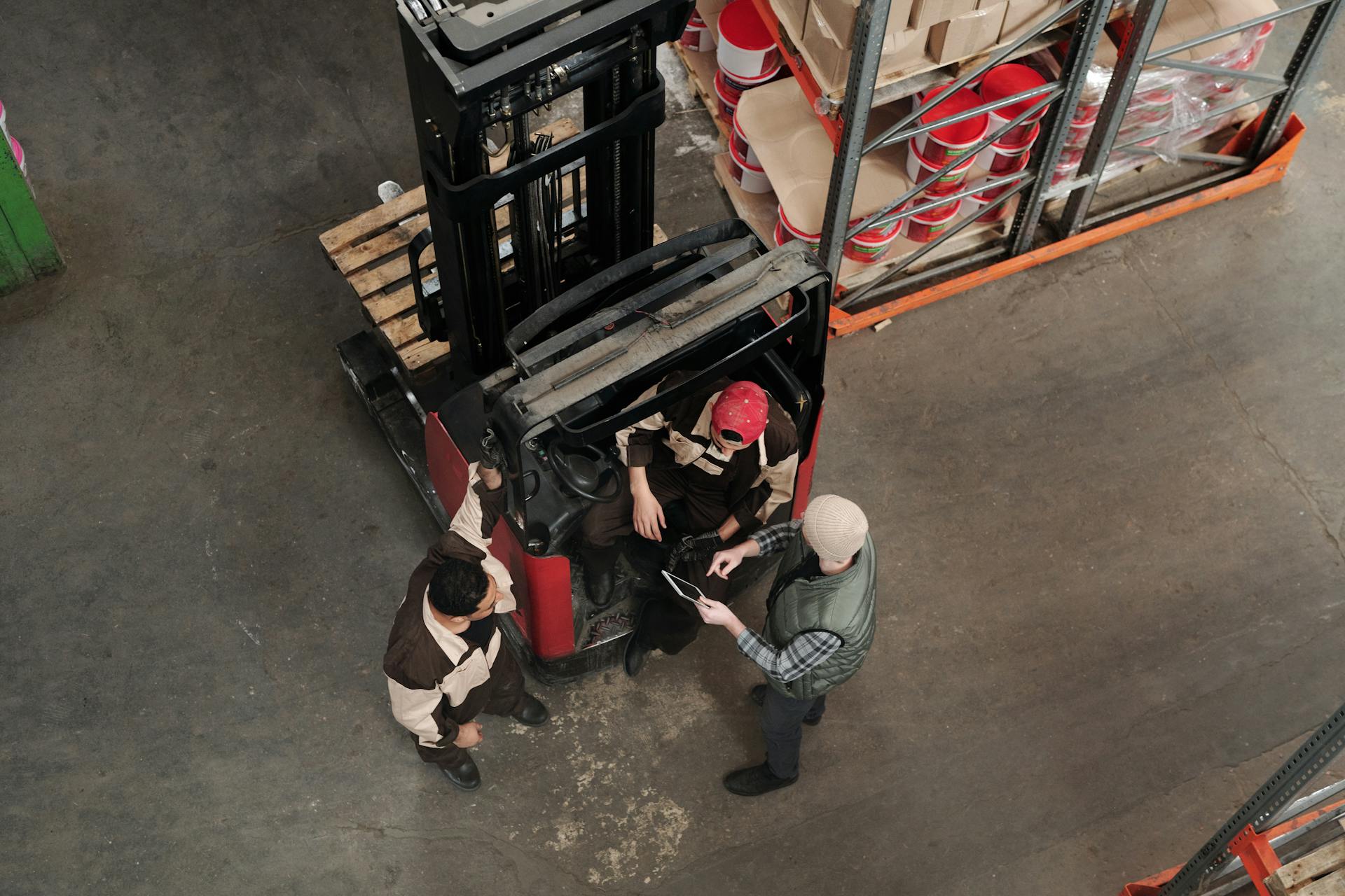 Top view of workers discussing plans in a warehouse beside a forklift, emphasizing teamwork.