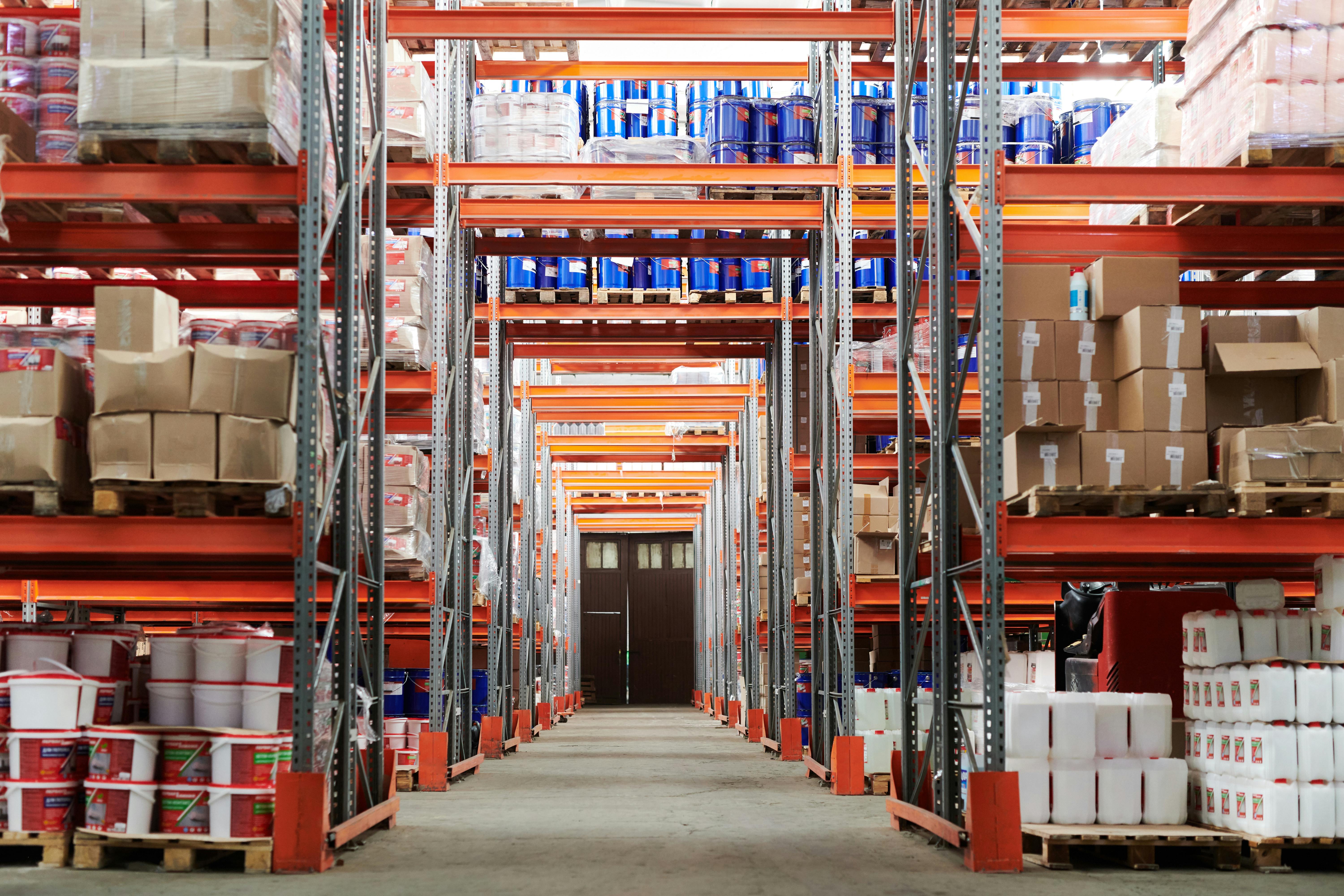 An image of the inside of a warehouse with various orange shelves with cardboard boxes on them. 