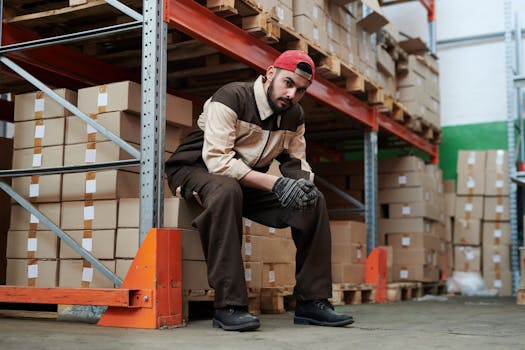 A warehouse worker in casual attire sits on a pallet amidst organized stacks of boxes.