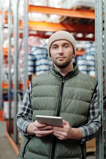A man wearing a beanie and vest holding a tablet in a warehouse setting. A man wearing a beanie and vest holding a tablet in a warehouse setting.