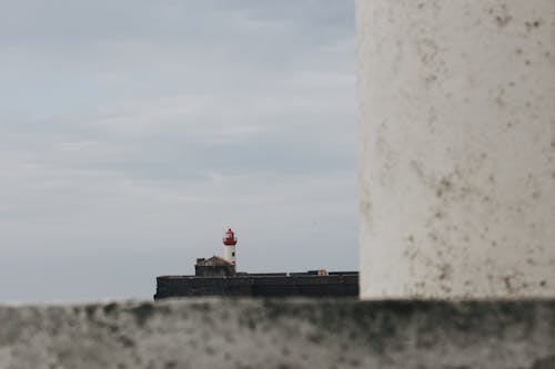 Distant lighthouse tower built on stony embankment seen from behind of concrete structure against overcast sky