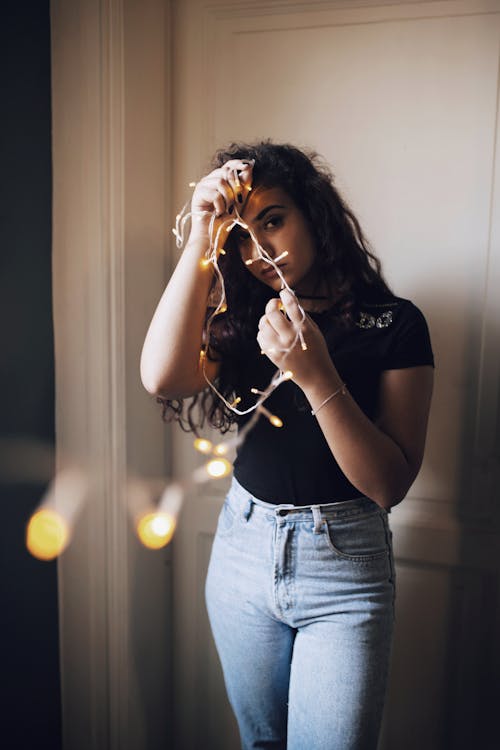 Young Hispanic female in casual outfit holding decorative light garland with glowing lamps and looking at camera while standing against door in room