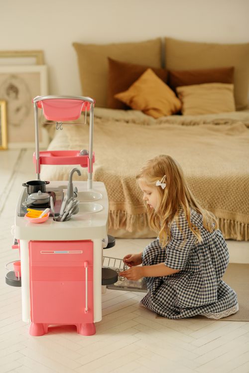 Free Adorable child in plaid dress playing with a toy kitchen indoors, capturing youthful imagination. Stock Photo
