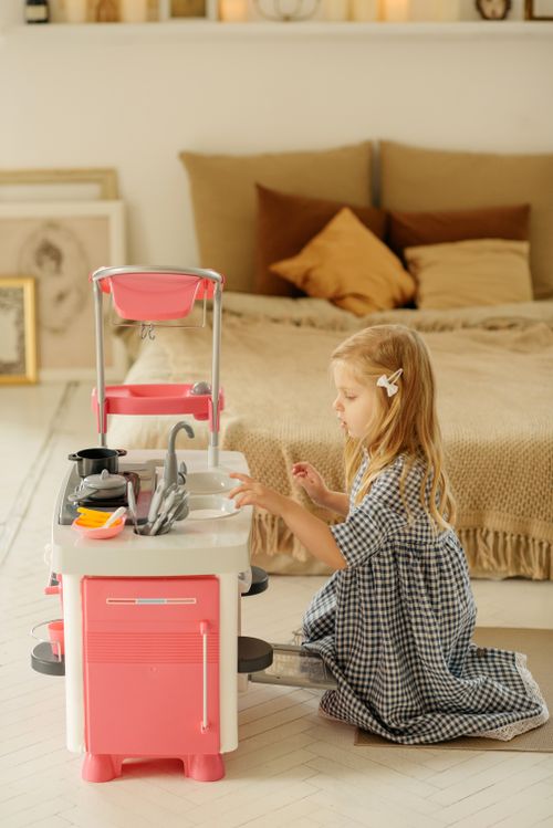 Free A blonde girl in a plaid dress playing with a toy kitchen set indoors, enjoying leisure time. Stock Photo