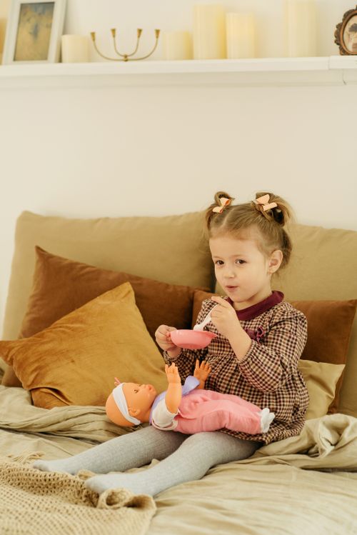 Free A young girl sits on a bed indoors, feeding a doll with a toy spoon. Stock Photo