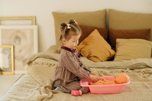 Free Girl in Plaid Dress Sitting on Bed While Playing With Baby Doll Stock Photo