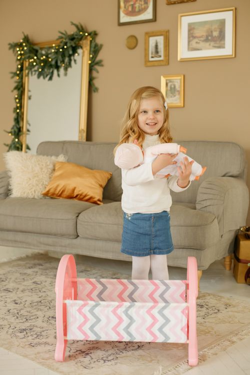 Free Joyful little girl standing in a living room, playing with a doll, surrounded by a festive atmosphere. Stock Photo