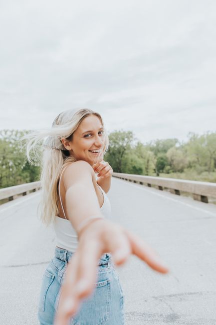 Cheerful young woman with blonde hair smiling on a scenic road, reaching out playfully.