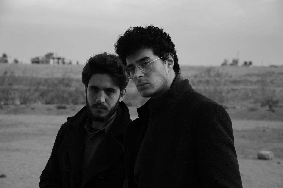 Black and white shot of serious young curly haired men in stylish clothes looking at camera while standing together on road against blurred field
