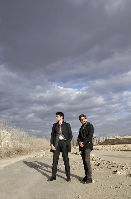 Full length of young men in classy warm outfits standing on sandy ground and looking at different directions against cloudy sky with urban buildings in background in sunny autumn day