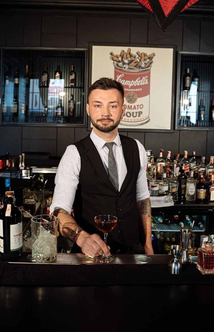 A Man In Black Vest Over White Long Sleeve Shirt Standing Inside Bar Counter Holding Glass Of Wine