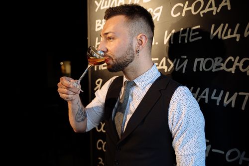 Free Elegant bartender in a suit tasting a cocktail with a backdrop of a bar's decorative text wall. Stock Photo
