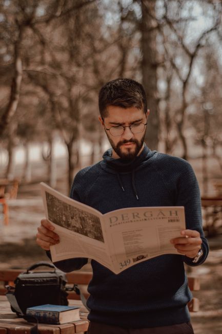 Young focused bearded ethnic male in eyeglasses reading magazine with illustration in city park Young focused bearded ethnic male in eyeglasses reading magazine with illustration in city park