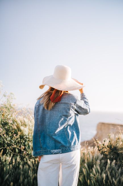 Woman in a denim jacket and wide-brimmed hat enjoying a sunny day by the sea.