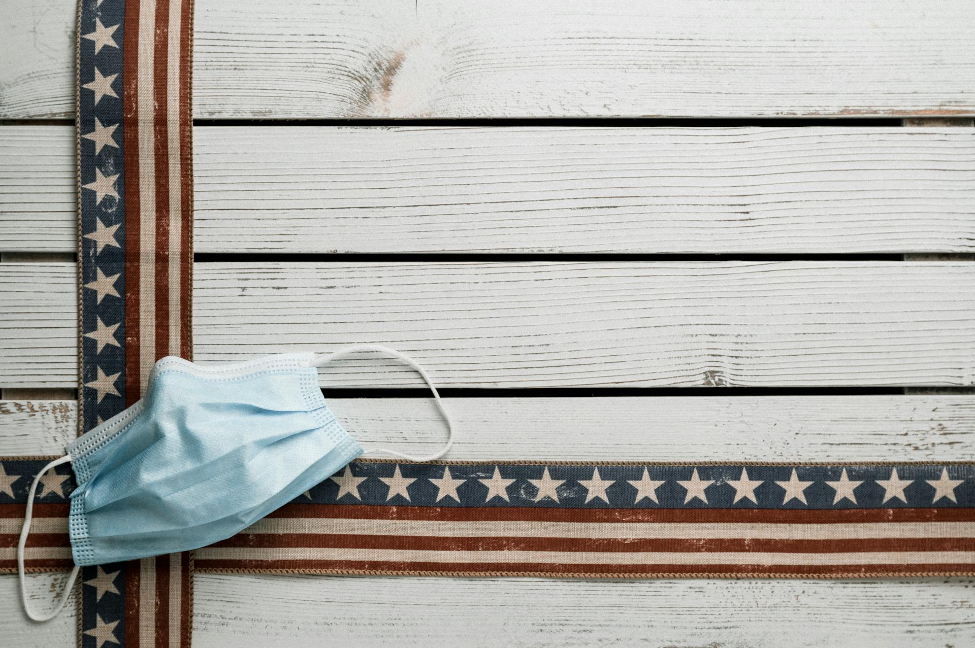 From above of sterile face mask on decorative USA ribbons with stripes and stars on wooden desk during COVID 19 pandemic