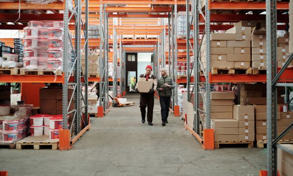 Storage Solutions โ Two workers handle a package in a spacious warehouse surrounded by shelves stocked with boxes and products.