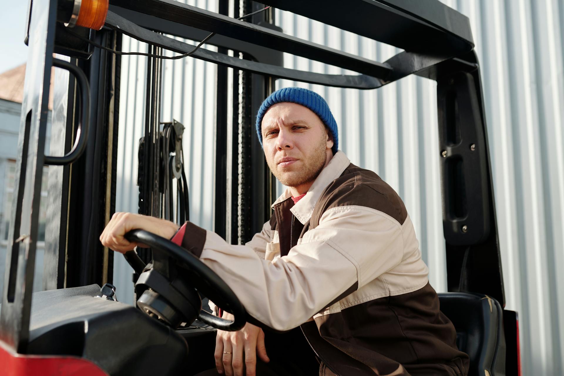 Male forklift operator wearing a blue beanie and work uniform, sitting outside on a sunny day.